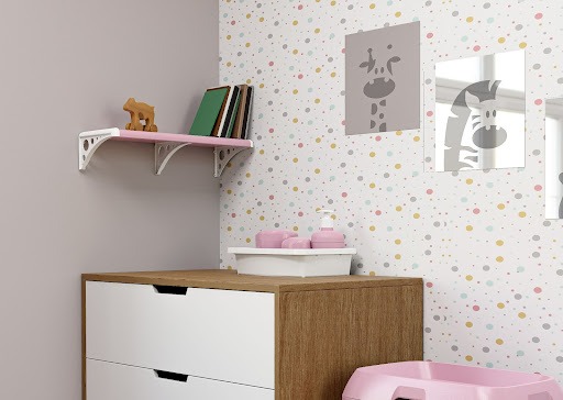  Um quarto infantil, com papel de parede de bolinhas coloridas que se localiza ao fundo do espaço. Uma cômoda acomoda um kit higiene da linha Astra Kids. Há uma prateleira rosa instalada na parede, com livros e um brinquedo sobre ela. Na parede ao fundo, há espelhos decorativos quadrados, com desenhos de girafa e zebra.