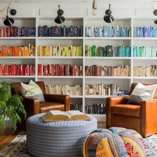 Uma grande estante, com livros organizados em degradê de cores, está em uma sala. A estante em madeira possui spots de luz acima. A sala possui um tapete no chão, junto a duas poltronas em couro e almofadas em cima. Dois puffs estão localizados ao centro, um com um livro aberto em cima. Ao lado, aparecem as folhagens de uma planta.