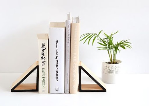  Uma mesa branca com quatro livros apoiados por aparadores de madeira em formato triangular, com as laterais pretas. Ao lado, há um vaso branco, com uma pequena planta.