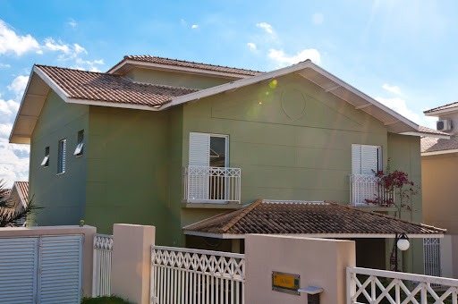 Casa de 2 andares, verde com telhado, à frente um muro com portão branco.