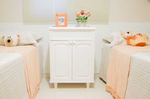 Quarto com duas camas de solteiro, ambas com lençol creme e manta laranja clara, mais ursos de pelúcia. Entre elas um gabinete plástico branco com um vaso de flores e porta-retratos acima. 