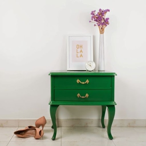 Na imagem está uma mesa de cabeceira com design antigo, pintada de verde, com um quadro branco, um relógio de mesa e vaso com flores de lavanda em cima. Ao lado, um par de sapatos de salto alto na cor nude. 
