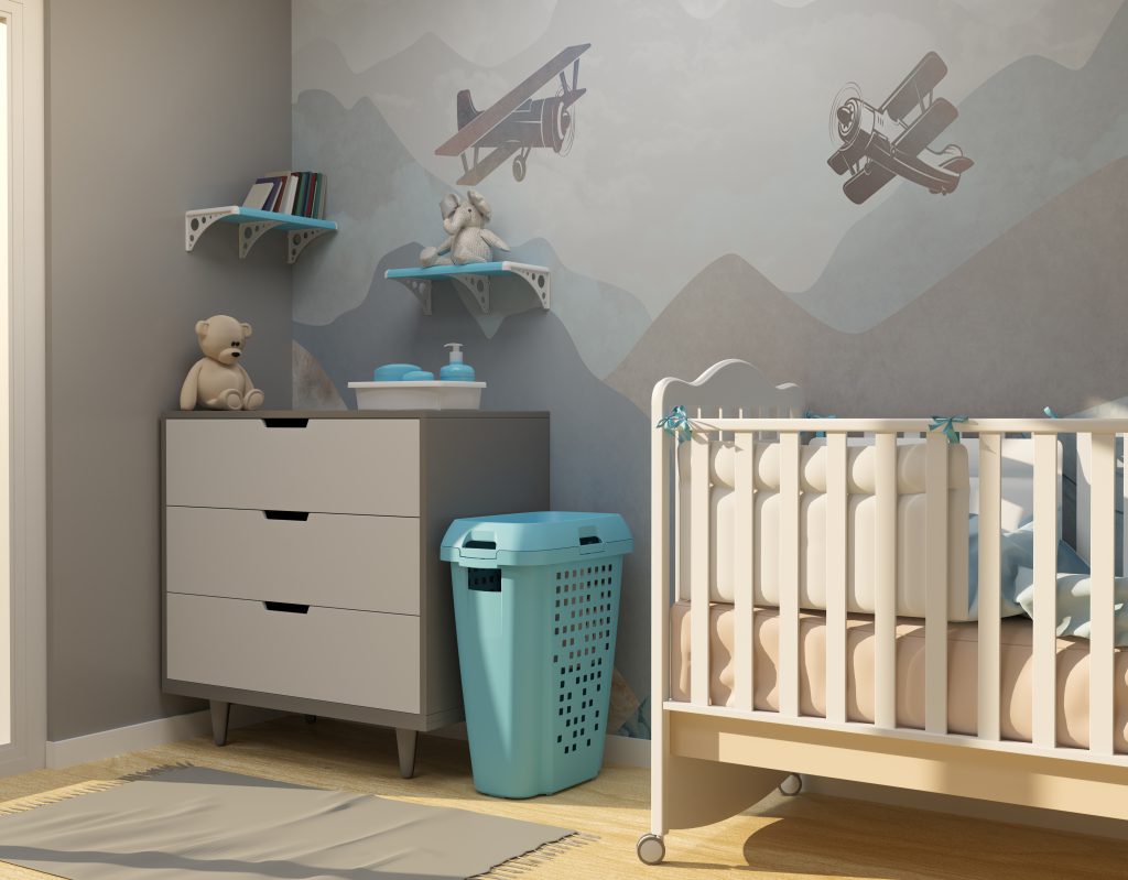 Quarto infantil com alguns itens da Astra em cor azul, com parede com desenhos de aviões e duas prateleiras plásticas. Em frente, um berço e uma cômoda, que possui um urso em cima, e ao lado, um roupeiro telado. 