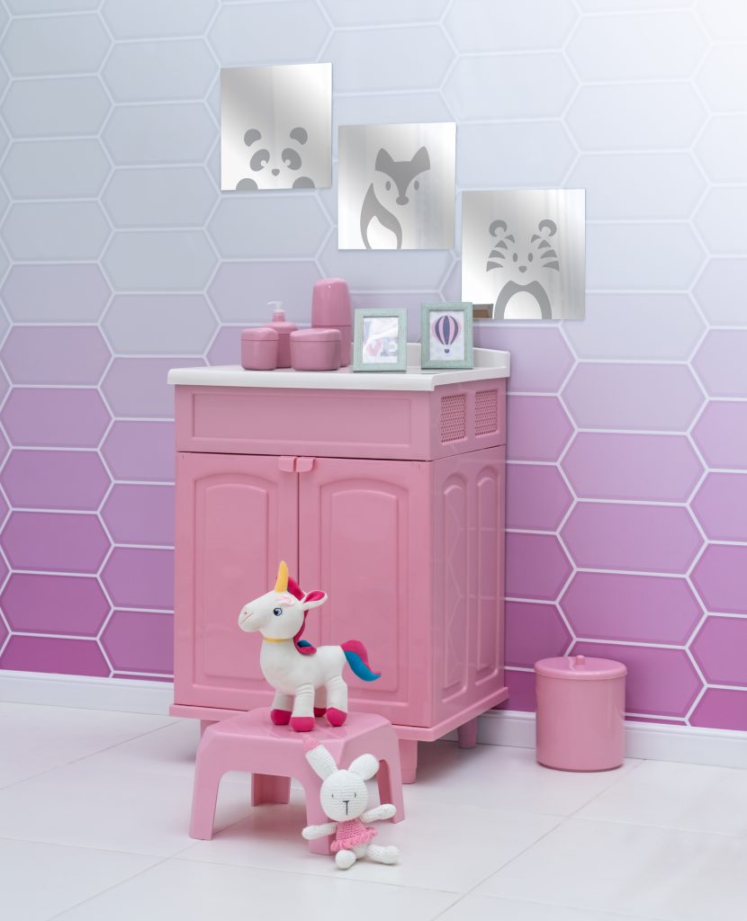 A imagem mostra um quarto em tons de rosa, com um gabinete plástico, porta-objetos e dois quadros em cima do móvel, lixeira e a mini banqueta da Astra, também na cor rosa. No espaço há pelúcias espalhadas, como um unicórnio e coelho. Na parede está aplicado um papel de parede com formatos hexagonais em degradê de rosa, além de espelhos decorativos com desenhos de bichos pendurados na parede.