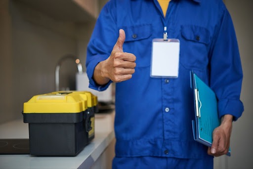 Na imagem temos um profissional com uma macacão azul, segurando uma prancheta e em cima da pia, temos a sua maleta de ferramentas, nas cores preta e amarela.