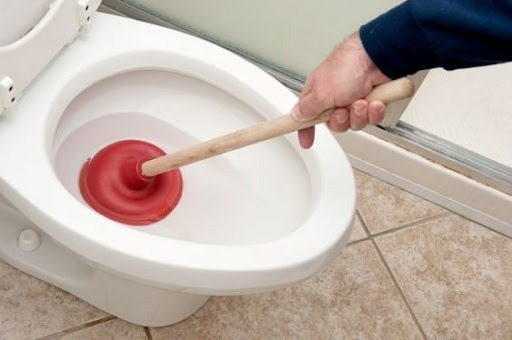 A imagem apresenta um banheiro com vaso sanitário branco ao lado do box do banheiro, e uma mão branca segurando um desentupidor. O item possui cabo de madeira e a base em borracha na cor vermelha. O vaso está com água dentro para facilitar o desentupimento. 