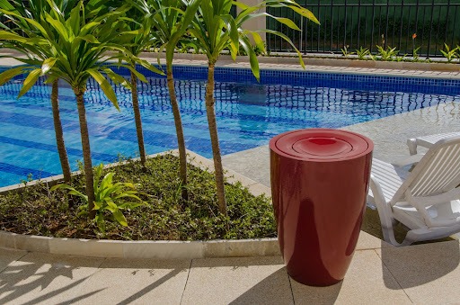 Em uma piscina de condomínio, temos a lixeira de fibra de vidro na cor vermelha à frente em destaque na imagem. ao lado esquerdo há um canteiro em formato triangular com plantas e alguns mini coqueiros, ao fundo aparece a piscina com água cristalina e ao lado esquerdo, atrás da lixeira, estão algumas espreguiçadeiras. 