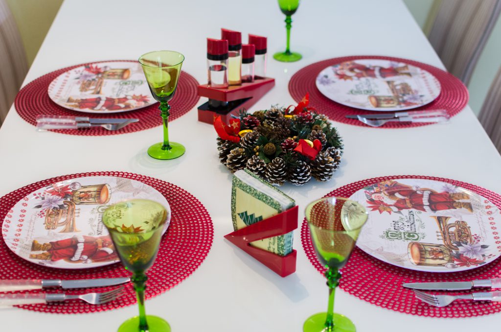 A imagem apresenta uma mesa de jantar na cor branca, decorada com itens natalinos, junto a pratos, guardanapos e um enfeite ao centro. Compõem a imagem o porta-guardanapos e o galheteiro da Astra, ambos na cor vermelha.