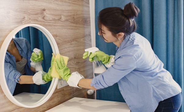 Na imagem uma mulher utilizando luvas, está realizando a limpeza de um espelho redondo. Ela está segurando um borrifador com alguma mistura e passando um pano no vidro do espelho.