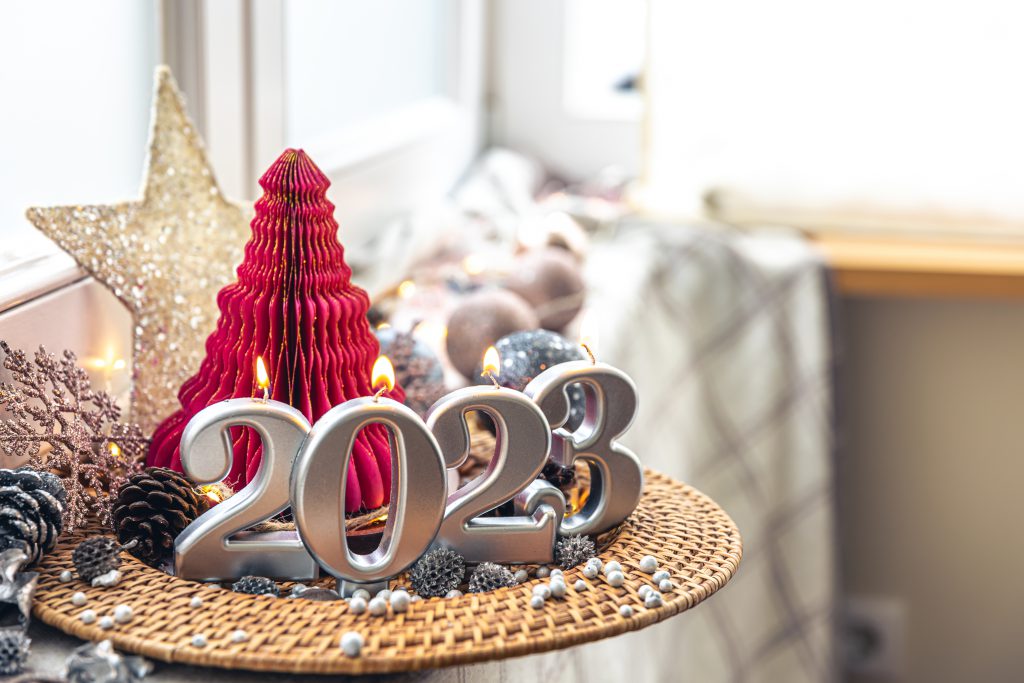 A imagem possui alguns itens de decoração para o Ano Novo. A frente um letreiro com o ano de 2023 na cor prata, ao fundo alguns itens como uma pequena árvore vermelha e uma estrela dourada.