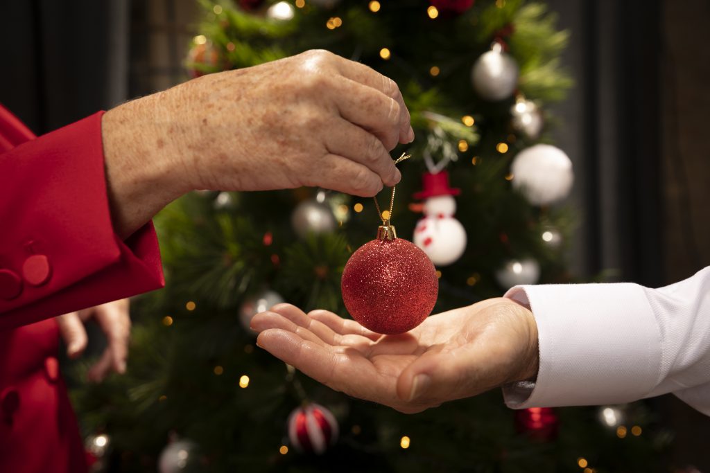 Na imagem temos duas mãos brancas, uma segurando um enfeite de bola e passando para a outra. Ao fundo vemos uma árvore de natal enfeitada, com as luzinhas ligadas. A imagem está com o fundo um pouco desfocado.