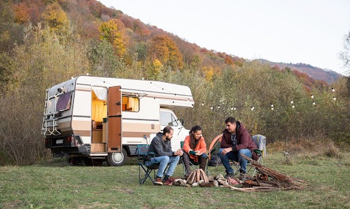 Na imagem temos um grupo de amigos fazendo uma fogueira ao ar livre em um ambiente aberto, sendo que logo atrás tem um motorhome branco com a porta aberta perto de árvores em tons de verde.