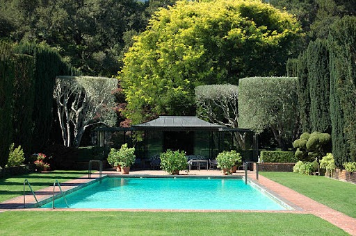 A imagem apresenta um grande jardim com área verde, com uma ampla piscina ao centro. A piscina é rodeada de grama e pequenos arbustos e ao fundo aparece uma casa com telhado que remete à arquitetura asiática. Atrás do espaço há árvores podadas em formatos e cores variadas. 