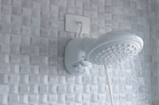 A imagem apresenta a parede de um banheiro e um chuveiro branco da Astra em funcionamento, com a água caindo do crivo. A parede atrás possui revestimento em pastilhas, e o acabamento da instalação é feito com um espelho de tomada também na cor branca. 