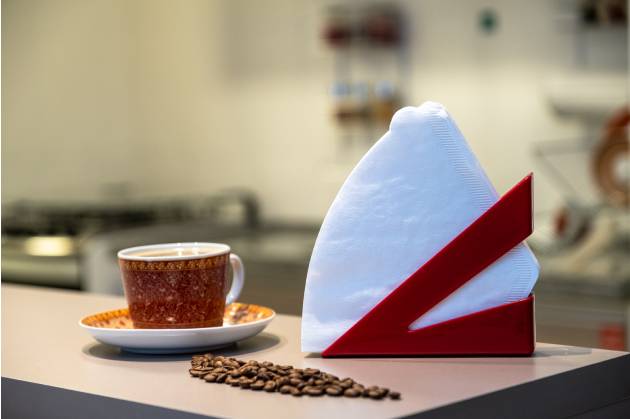  A imagem mostra uma bancada de uma cozinha americana de apartamento, neste espaço estão grãos de café espelhados ao lado de um porta-guardanapo vermelho com filtro de papel branco próximo a um pires e uma xícara de café.