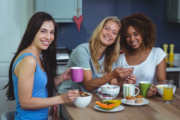 A imagem mostra um grupo de três amigas felizes tomando café da manhã. Elas estão lado a lado em uma  mesa cheia de comidas de café da manhã, como bolinhos, mamão, cereal, sucos e café em xícaras.