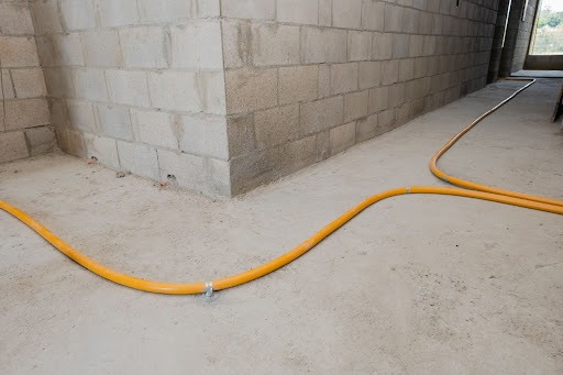 Em uma obra, aparecem paredes de tijolos de concreto à mostra. O chão, ainda no concreto, apresenta uma tubulação flexível para gás, na cor amarela. A imagem apresenta dois tubos fazendo curvas, em uma bifurcação para a esquerda e outra para a direita.  