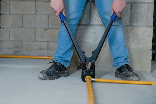 Em uma obra, parte das pernas de um homem branco de calça jeans azul e botinas pretas aparece fazendo a instalação de tubos flexíveis para gás, na cor amarela, com um alicate específico para este trabalho. O espaço está com as paredes de tijolos e chão ainda sem acabamento. 