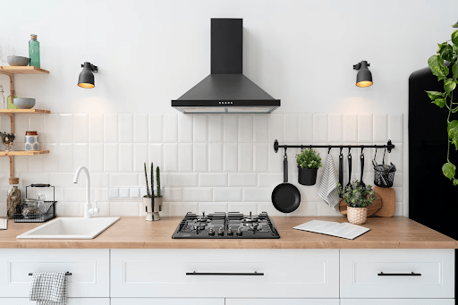 A imagem possui uma bancada de cozinha, em estilo escandinavo, com um fogão embutido preto e uma torneira branca. Logo acima, está instalado uma coifa/exaustor preto e alguns acessórios pendurados na parede. 