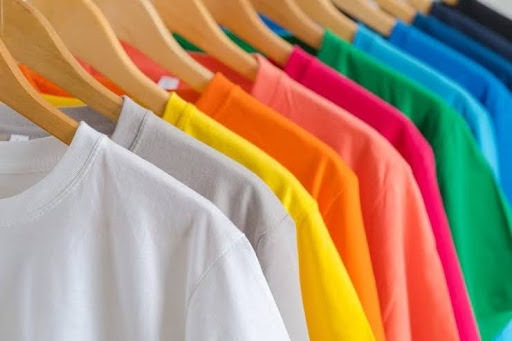 A imagem apresenta várias camisetas coloridas penduradas em cabides, feitos de madeira.