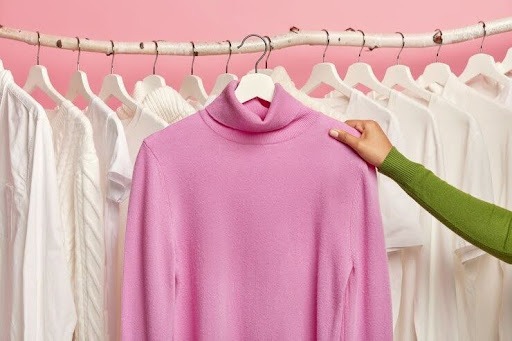 A imagem mostra um braço com uma blusa de manga verde segurando uma blusa rosa, e atrás estão várias camisetas brancas penduradas em uma espécie de arara de roupas. 