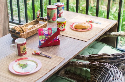 A imagem mostra uma mesa de madeira em um ambiente externo onde que em cima da mesa temos um cesto de trançado com palha com alguns pães em seu interior, dois potes com decorações natalinas, duas canecas natalinas,, dois pratos com decoração de natal, duas colheres e um porta guardanapos na cor vermelha.