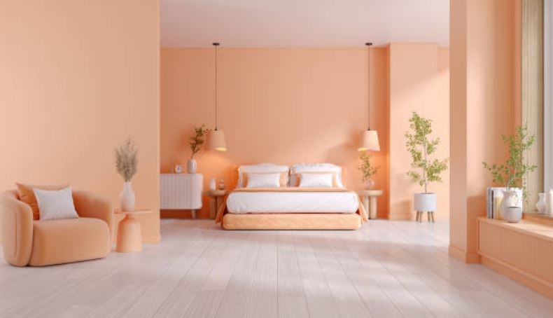 Quarto aconchegante em tons de pêssego (Peach Fuzz), a cor do ano da Pantone. A cama de casal com lençol branco e colcha bege convida ao relaxamento. Um criado-mudo de madeira clara com um abajur branco e uma planta verde em um vaso completam o visual. Uma grande janela com cortinas brancas abertas deixa entrar a luz natural, enquanto uma mesa de cabeceira de madeira clara com uma luminária pendente e um tapete bege felpudo no chão criam um ambiente acolhedor.