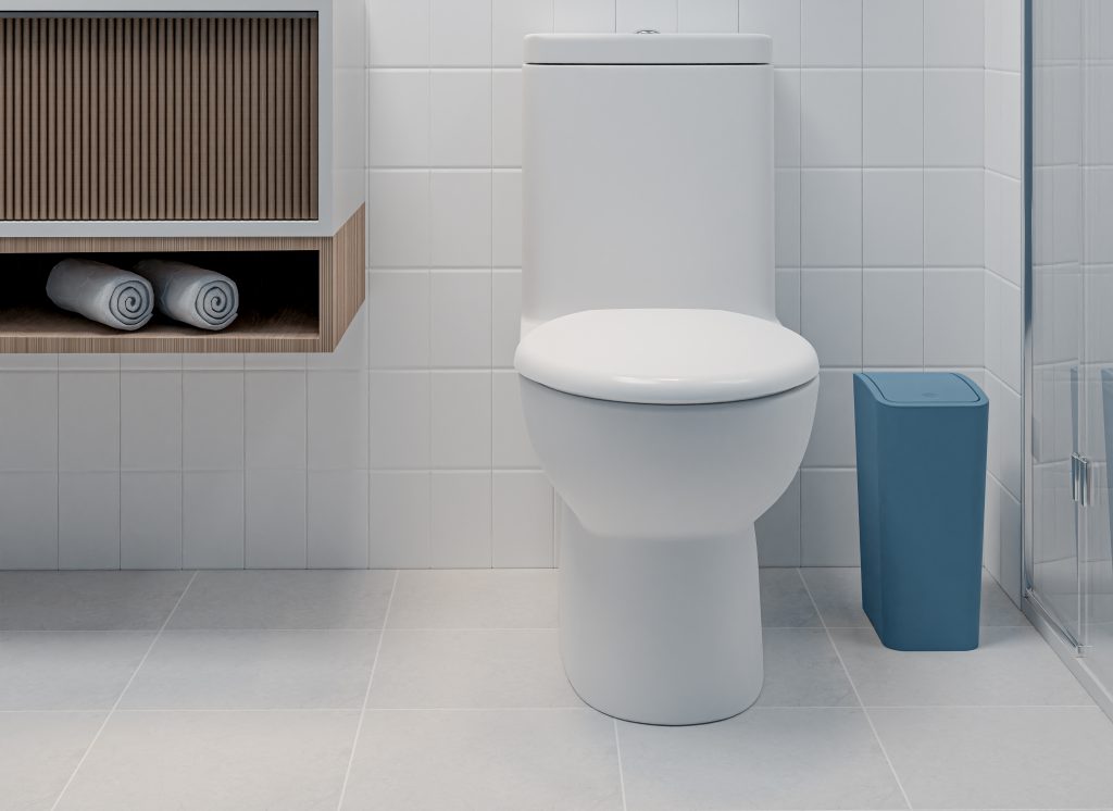 Um vaso sanitário branco ao lado de um cesto de lixo na cor mediterrânea em um banheiro simples. O piso do banheiro é cinza e as paredes são brancas. Há uma parte de um gabinete de madeira com uma cuba de apoio branca no canto superior esquerdo da imagem.