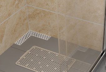 Parte de um banheiro aparece na imagem. Da esquerda para a direita, há a parte do box, com piso cinza, um ralo de canto e tapete de banho. Do lado de fora do box, há um vaso sanitário com caixa acoplada branco e assento sanitário bege. 